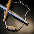 a necklace made with a tan coloured suede cord on which a piece of drumstick has been added - the drumstick has a circle pattern etched into it - the necklace rests on a pair of drumsticks
