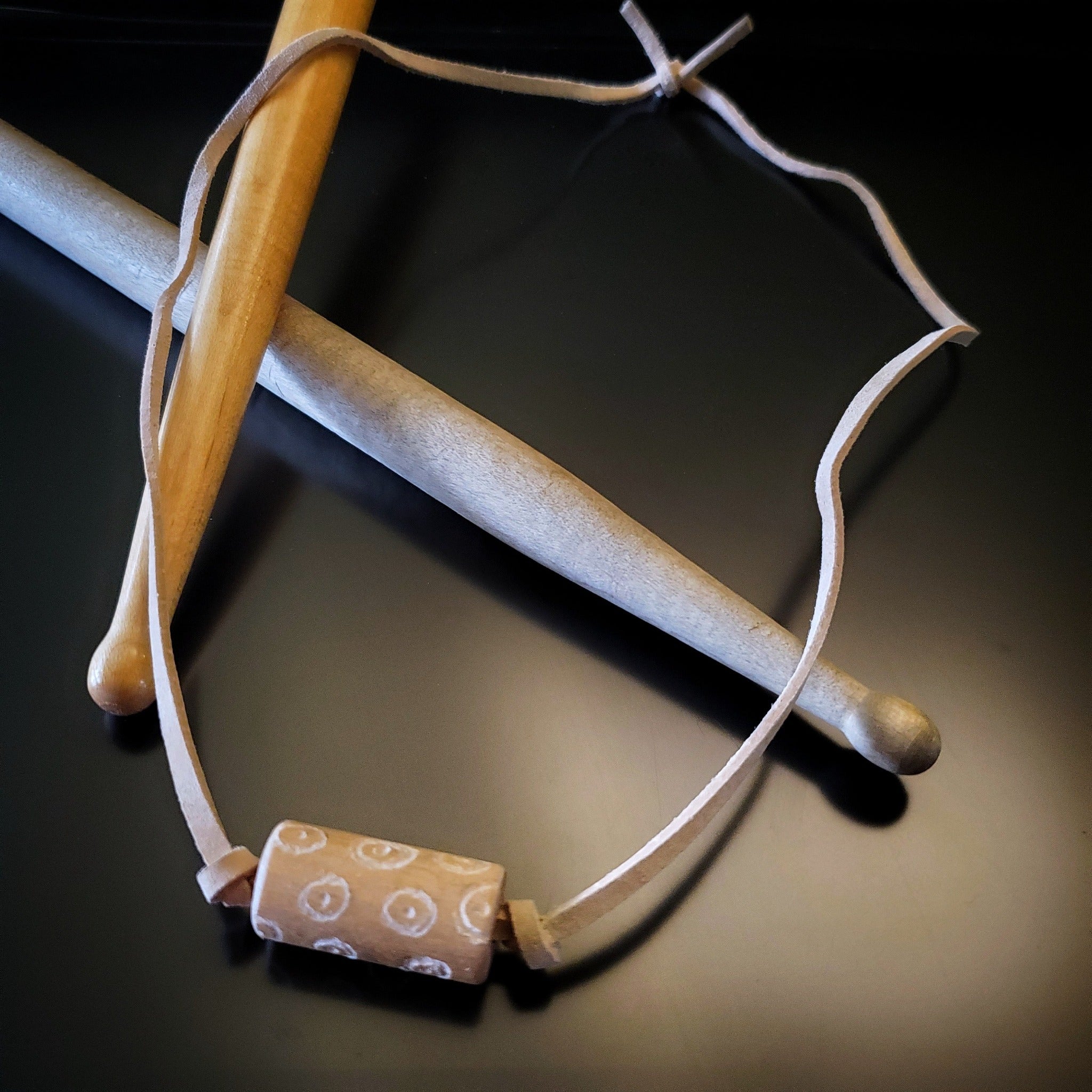 a necklace made with a tan coloured suede cord on which a piece of drumstick has been added - the drumstick has a circle pattern etched into it - the necklace rests on a pair of drumsticks