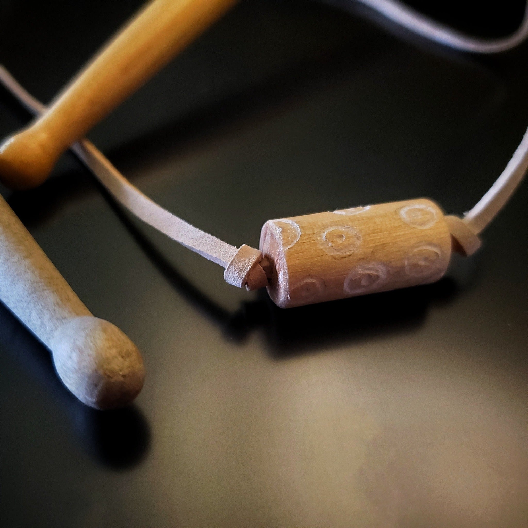 close-up of a a necklace made with a tan coloured suede cord on which a piece of drumstick has been added - the drumstick has a circle pattern etched into it - the necklace rests on a pair of drumsticks