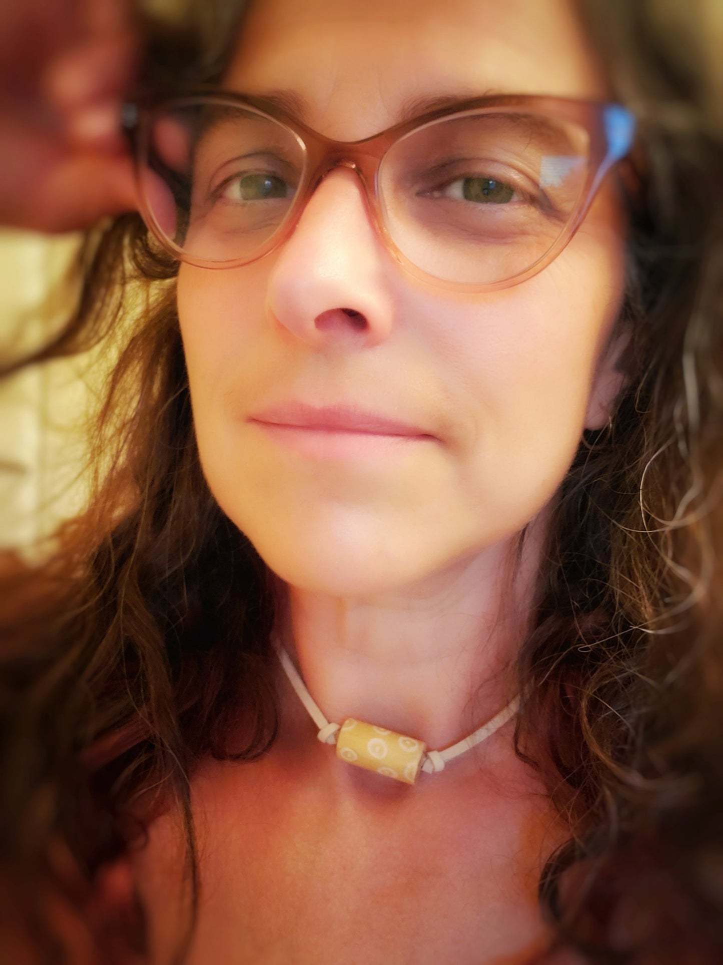 woman's head wearing glasses and long curly brown hair- she is wearing a a necklace made with a tan coloured suede cord on which a piece of drumstick has been added - the drumstick has a circle pattern etched into it 
