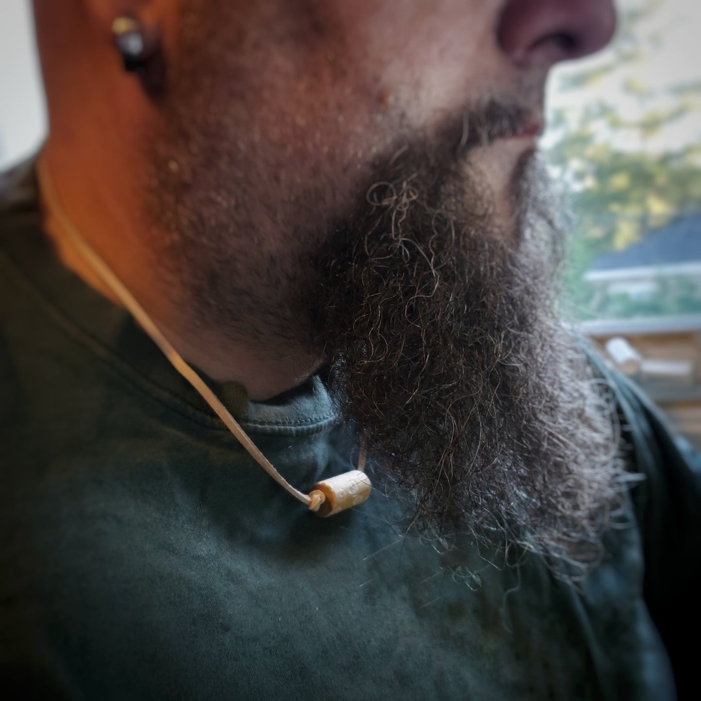 bottom of a man's head / profile (he has a beard and mustache) - he is wearing a a necklace made with a tan coloured suede cord on which a piece of drumstick has been added - the drumstick has a circle pattern etched into it 