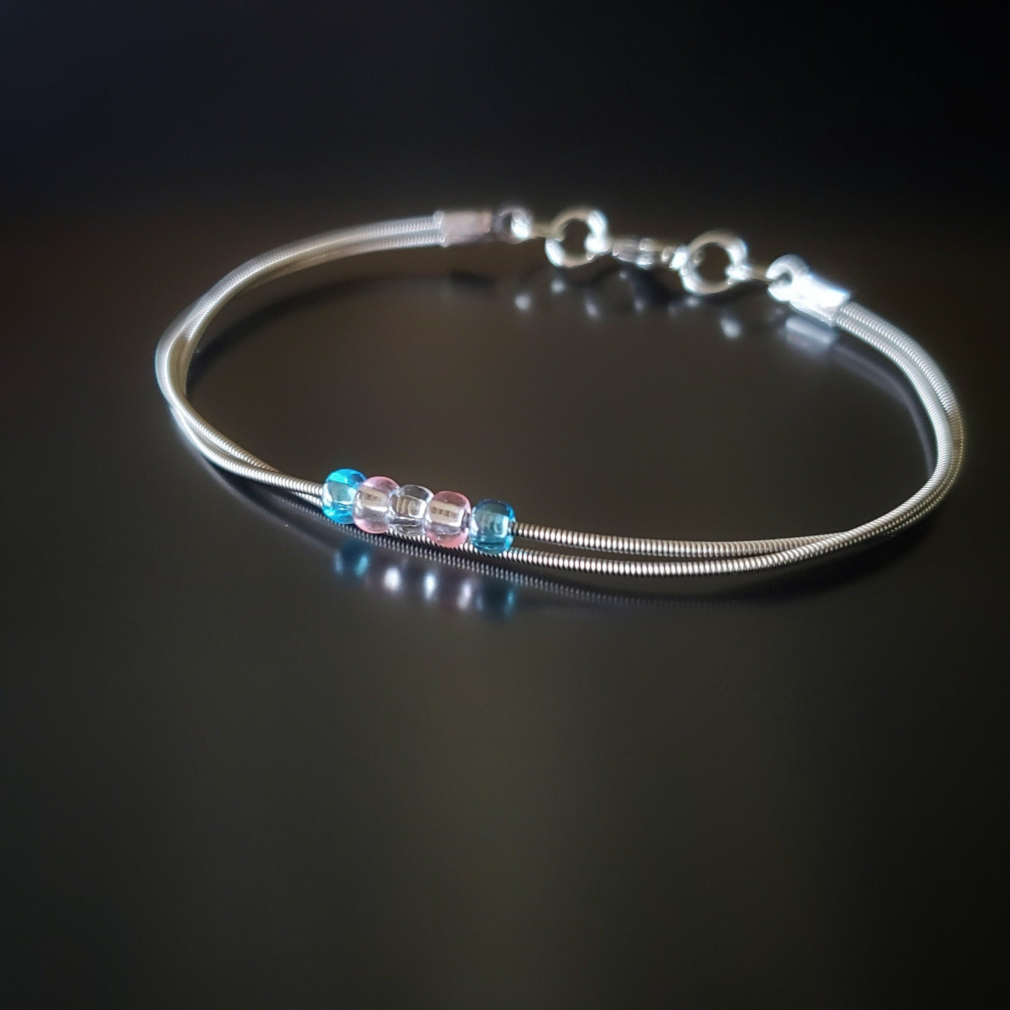 silver coloured clasp style bracelet made from upcycled guitar strings - there are 5 beads representing the colours of the transgender pride flag - 2 blue, 2 pink and 1 white