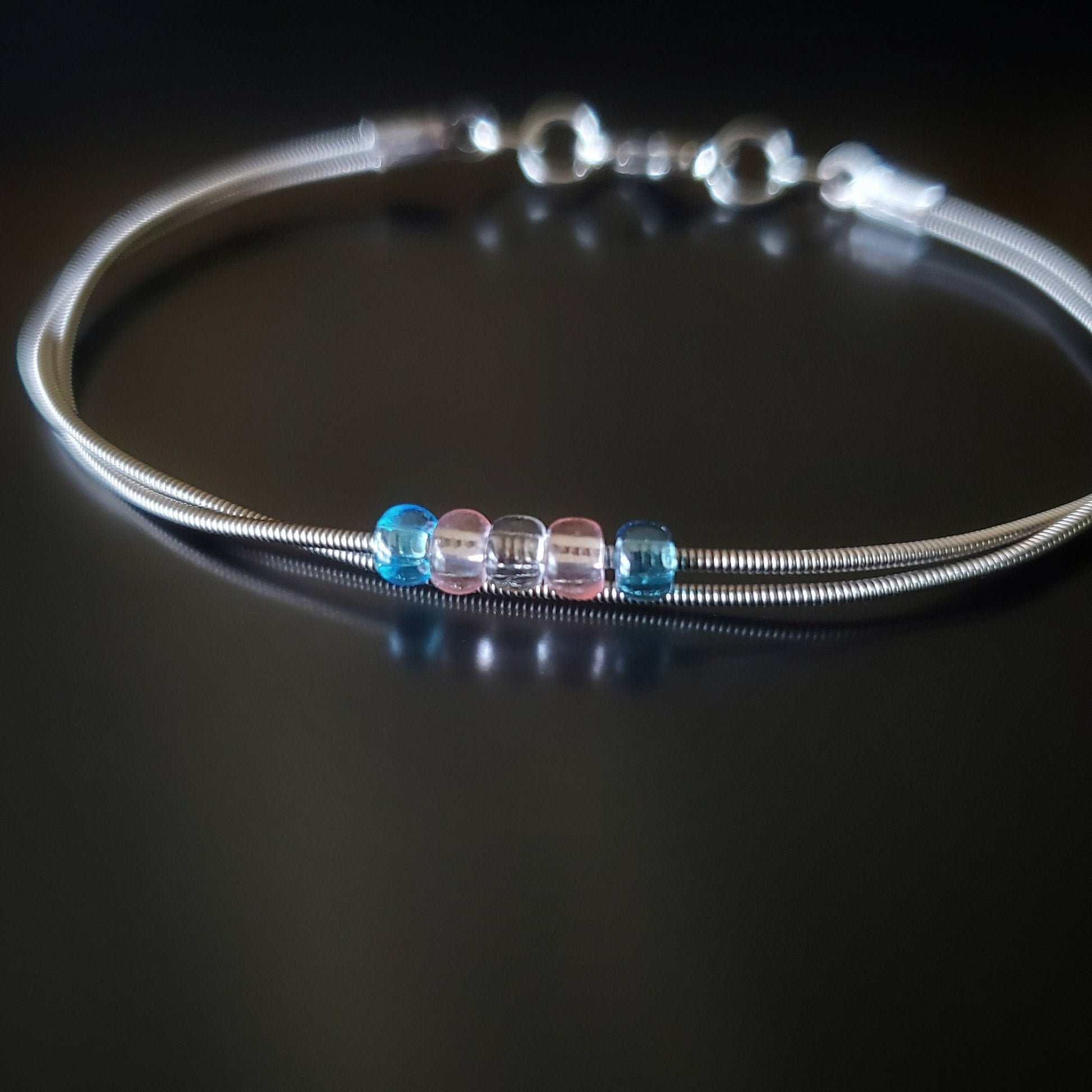 silver coloured clasp style bracelet made from upcycled guitar strings - there are 5 beads representing the colours of the transgender pride flag - 2 blue, 2 pink and 1 white
