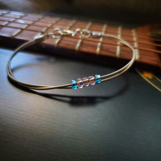 silver coloured clasp style bracelet made from upcycled guitar strings - there are 5 beads representing the colours of the transgender pride flag - 2 blue, 2 pink and 1 white- bracelet is sitting on the neck of a black guitar