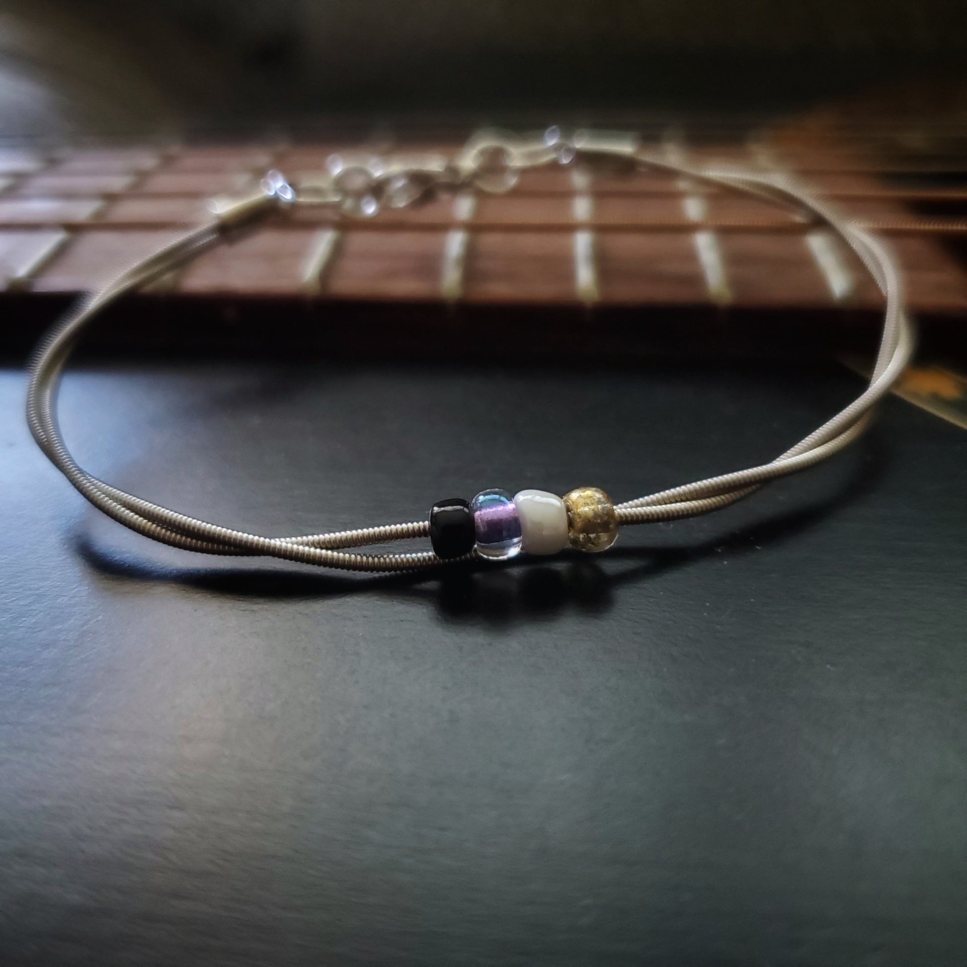 silver coloures clasp style bracelet with 4 glass beads representing the non-binary flag (yellow, white, purple & black) -bracelet is in front of a guitar neck that has no strings