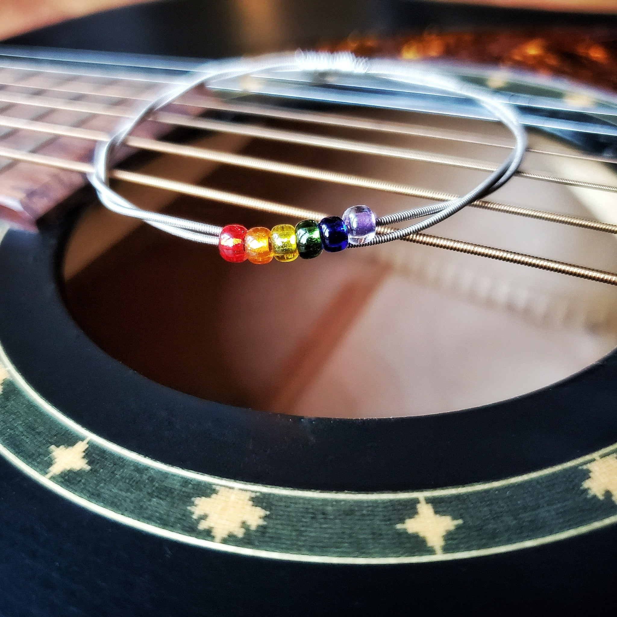 bangle style bracelet made from an upcycled guitar string - 6 glass beads represent the colours of the LGBTQ flag (red, orange, yellow, green, blue and purple) - sitting on the strings of a black guitar
