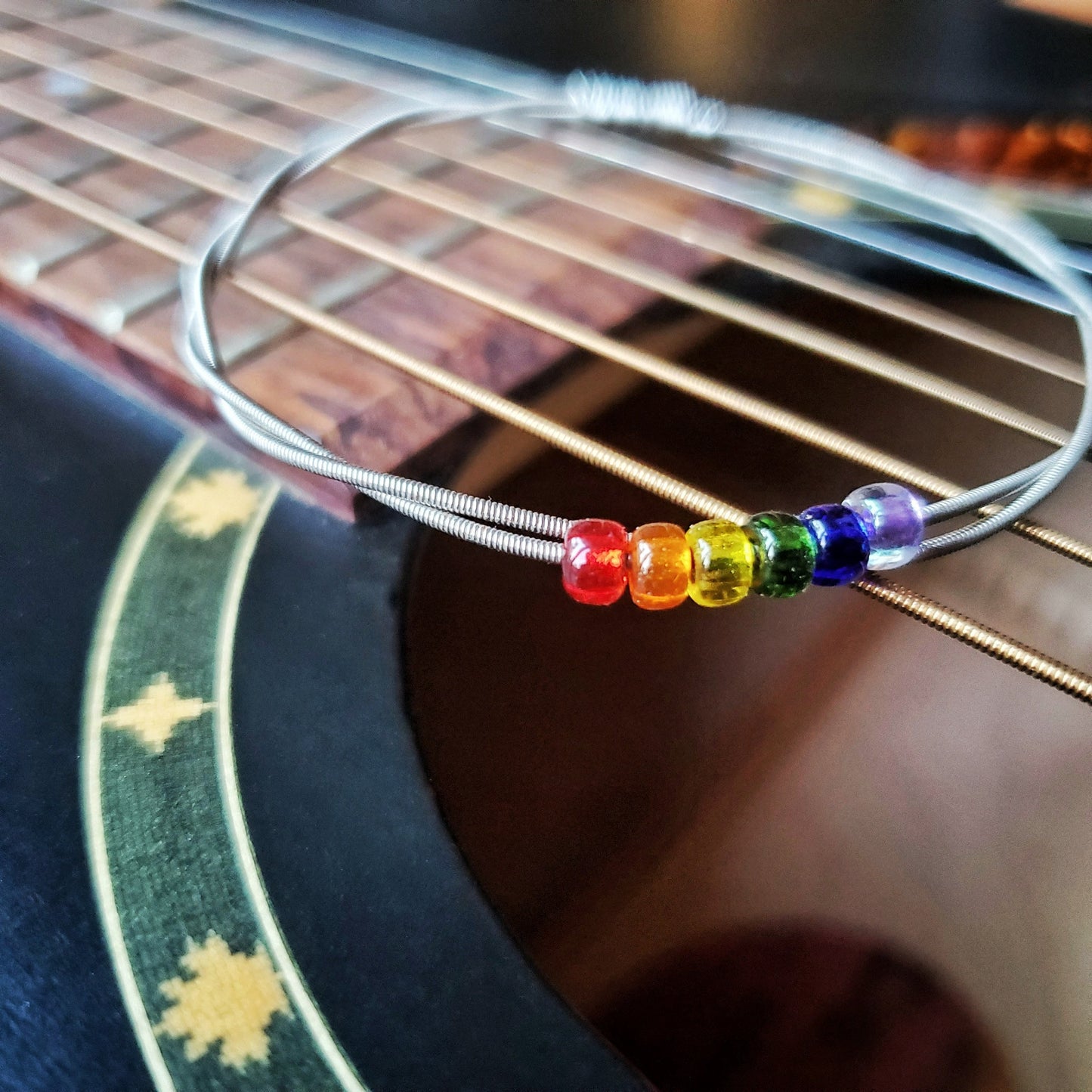 bangle style bracelet made from an upcycled guitar string - 6 glass beads represent the colours of the LGBTQ flag (red, orange, yellow, green, blue and purple) - sitting on the strings of a black guitar