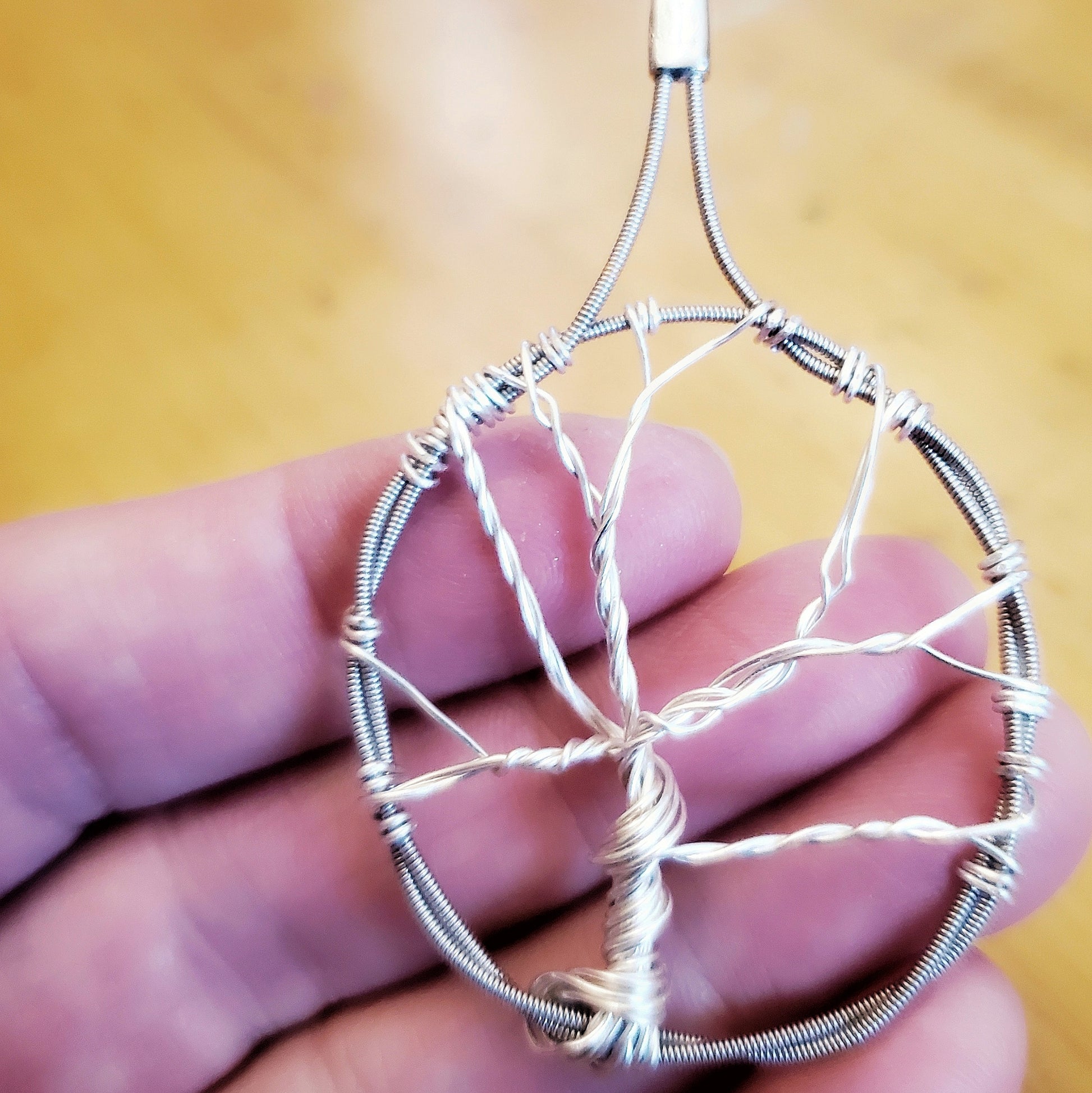 a pendant in the shape of a tree with a circle around it - the tree is made of silver coloured wire and the circle is made from upcycled guitar strings - the pendant sits on a person's fingers