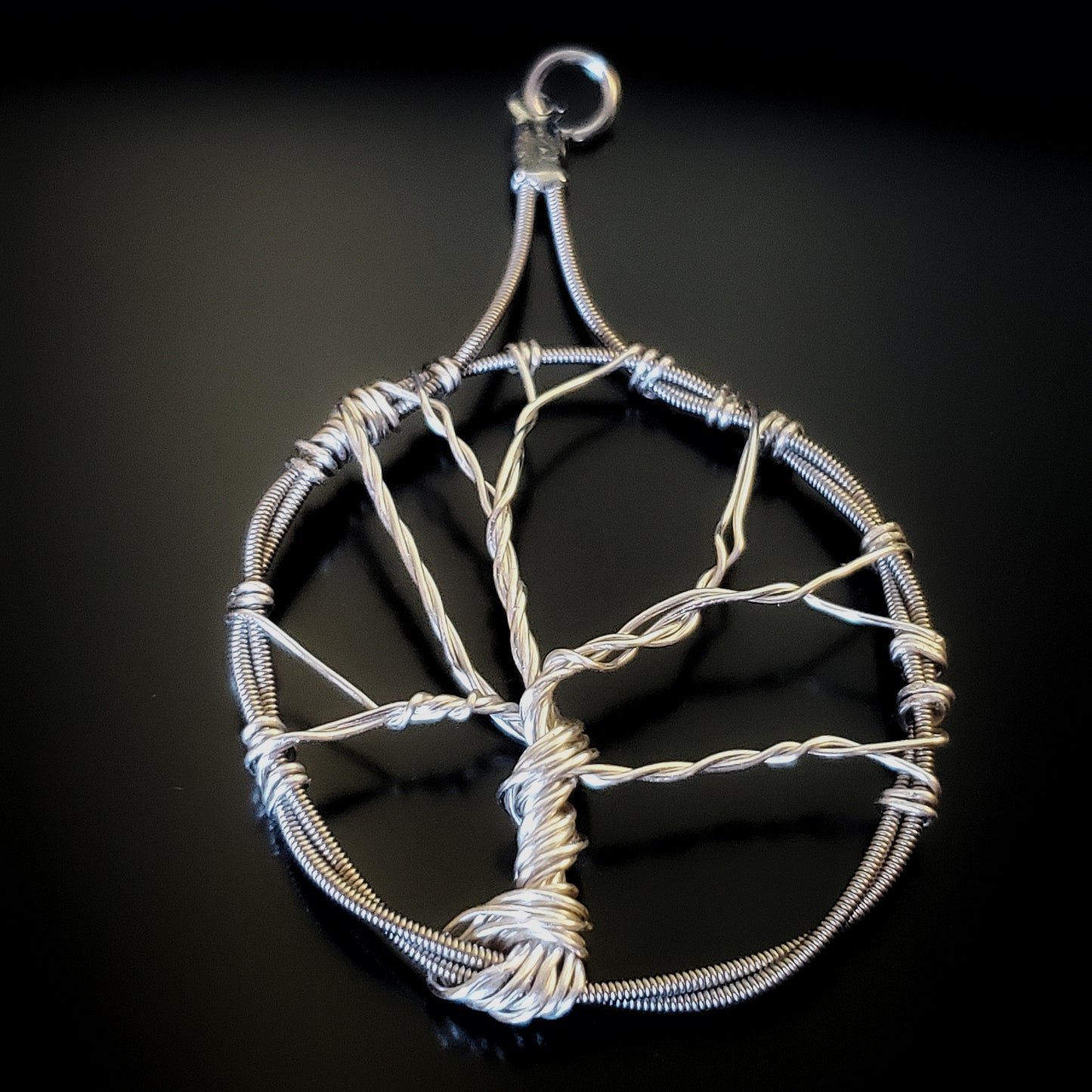 a pendant in the shape of a tree with a circle around it - the tree is made of silver coloured wire and the circle is made from upcycled guitar strings - black background