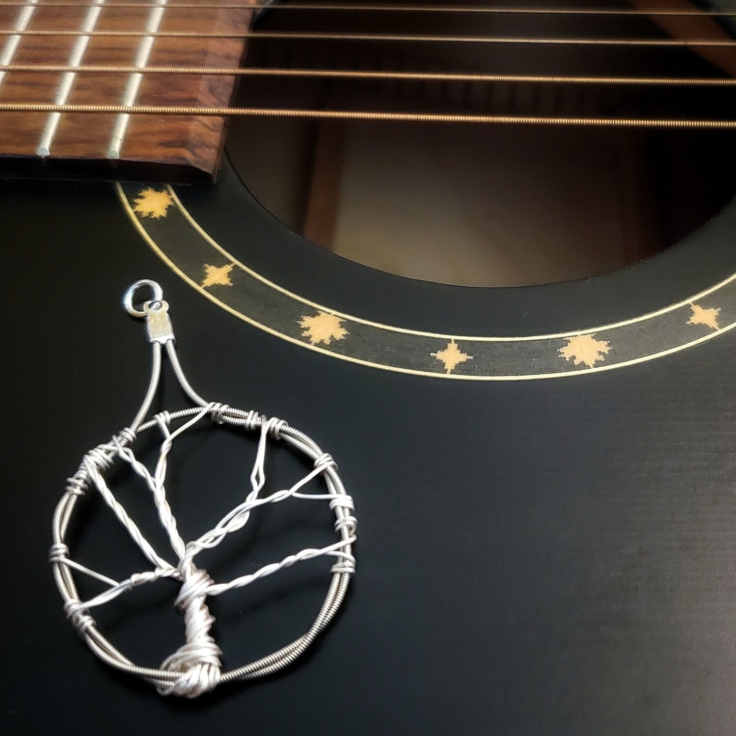 a pendant in the shape of a tree with a circle around it - the tree is made of silver coloured wire and the circle is made from upcycled guitar strings - the pendant lies the body on a black guitar