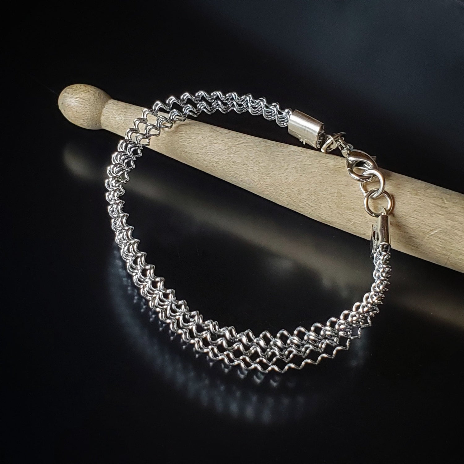 silver coloured clasp style bracelet made from a series of upcycled snare drum strings - bracelet is lying across a drumstick