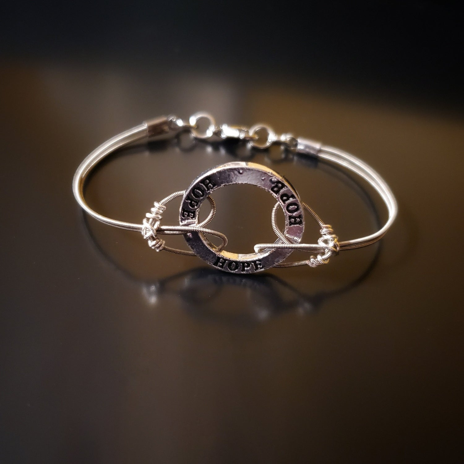 bracelet made from upcycled guitar strings connected to a silver hoop shaped charm with the word HOPE etched 3x on it
