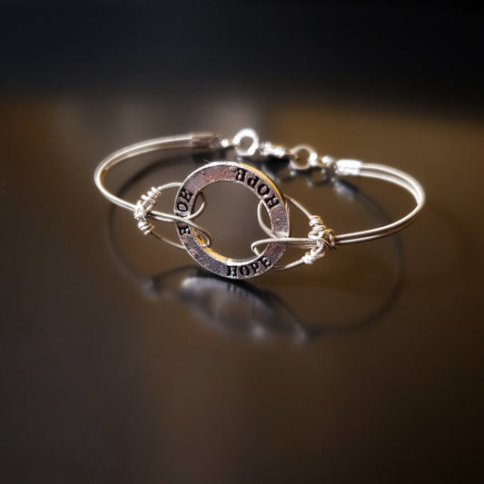 bracelet made from upcycled guitar strings connected to a silver hoop shaped charm with the word HOPE etched 3x on it