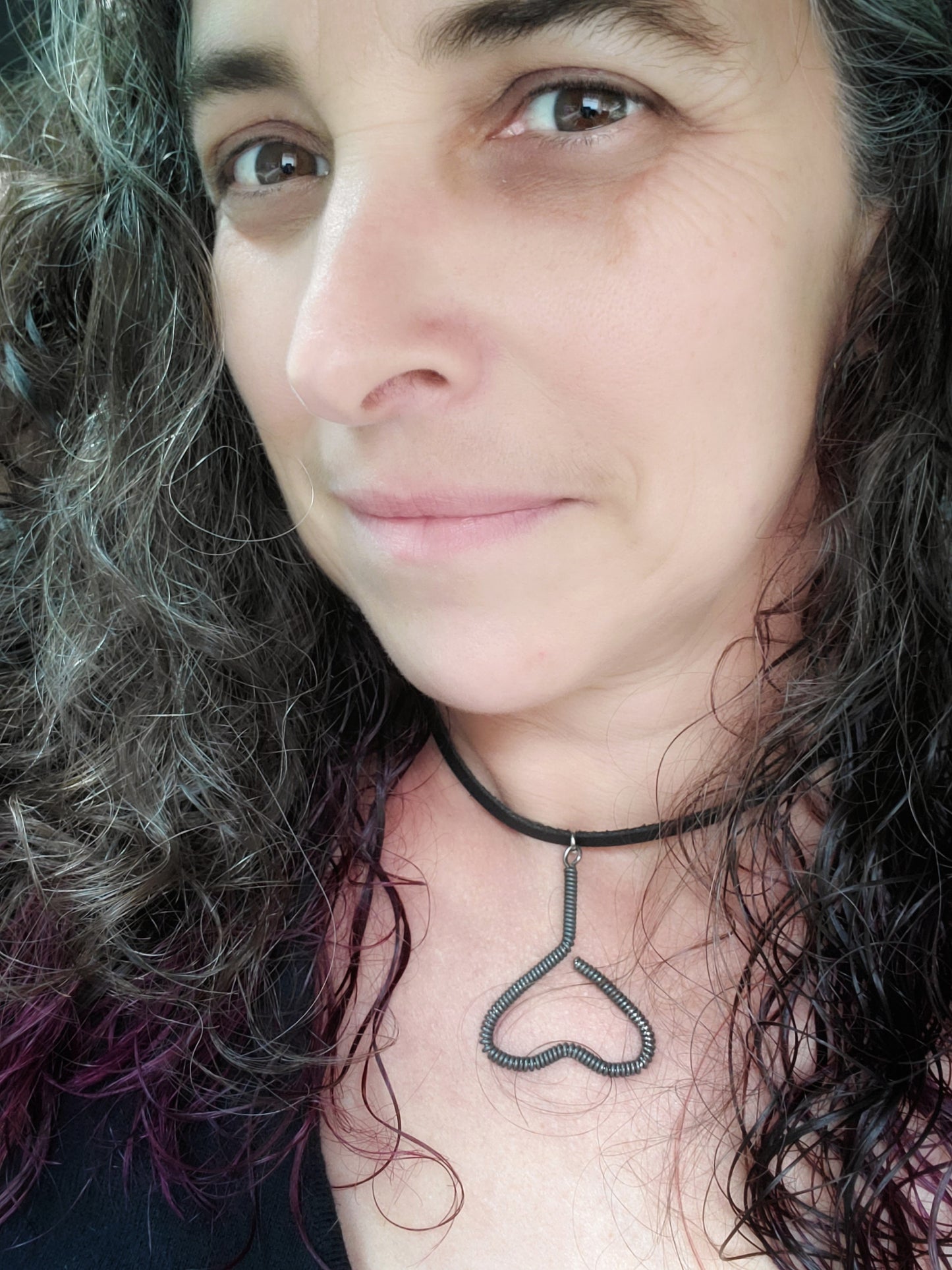 woman's head with black, grey and purple hair - she is wearing a necklace - pendant is in the shape of an upside down heart and is made from a black upcycled piano string - pendant hangs off a black leather cord