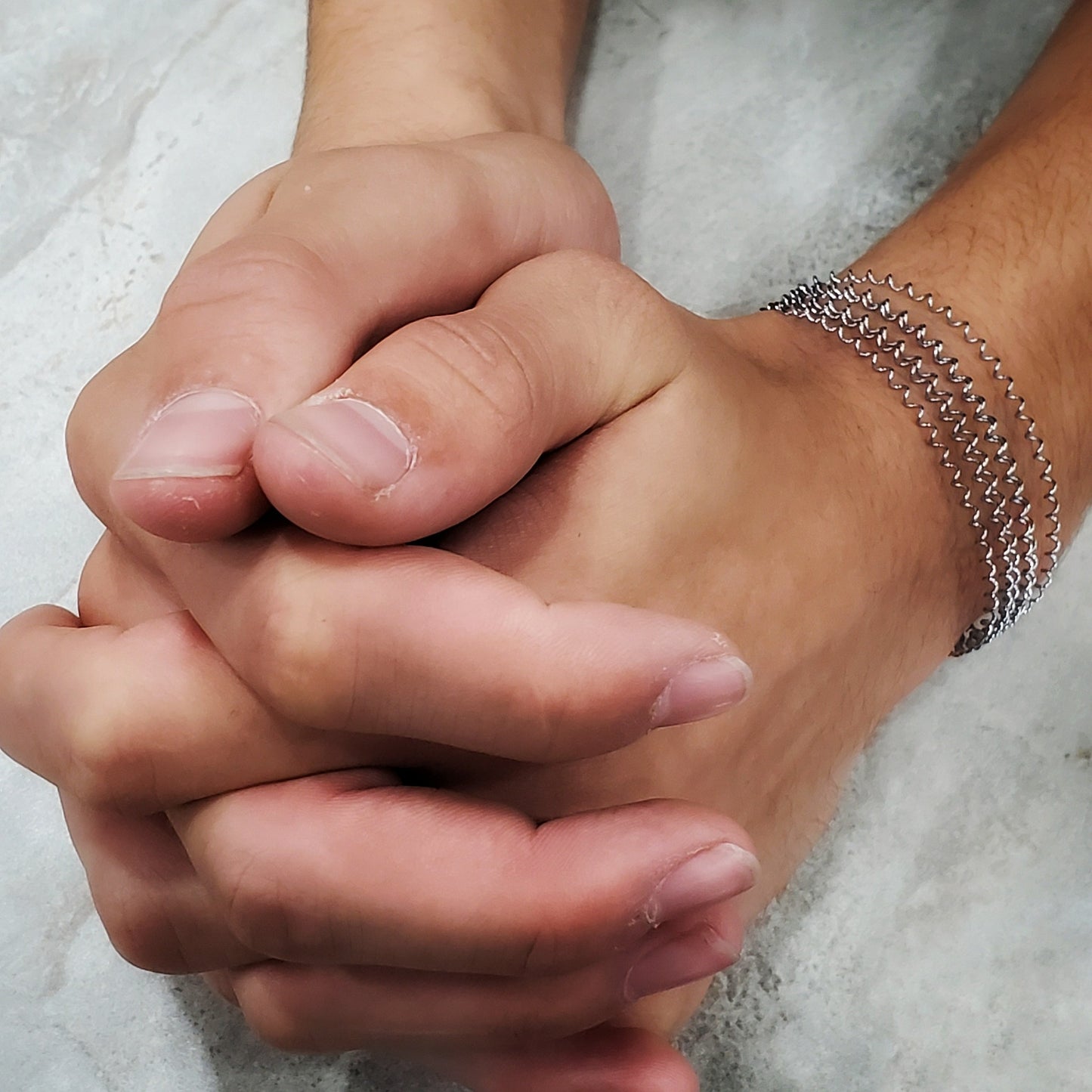 two male hands with the fingers intertwined on one wrist he is wearing a silver coloured clasp style bracelet made from a series of upcycled snare drum strings