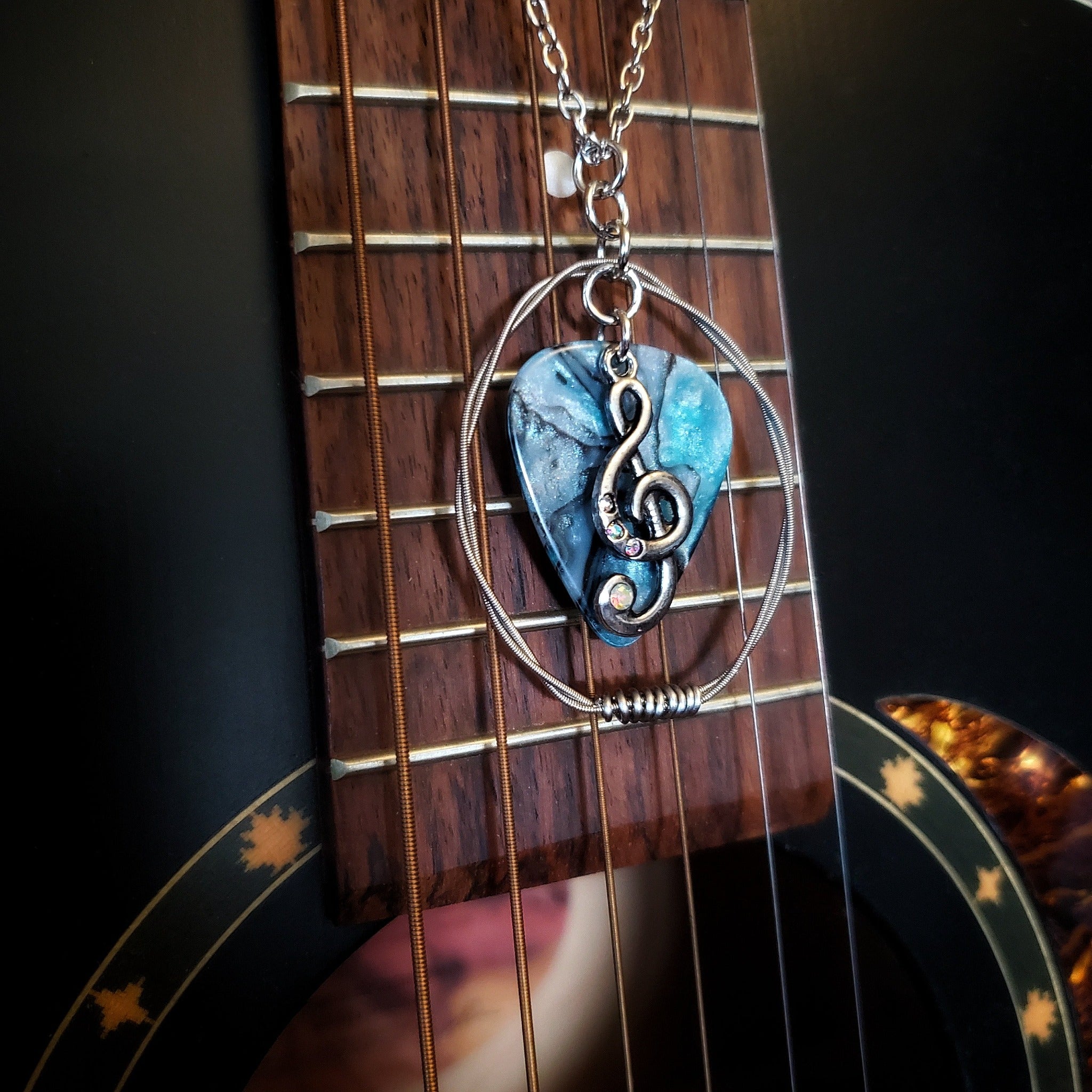 A necklace made from a blue marbled guitar pick and upcycled guitar strings with a silver coloured chain hangs in front of a black guitar