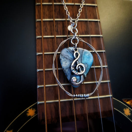 A necklace made from a blue marbled guitar pick and upcycled guitar strings with a silver coloured chain hangs in front of a black guitar