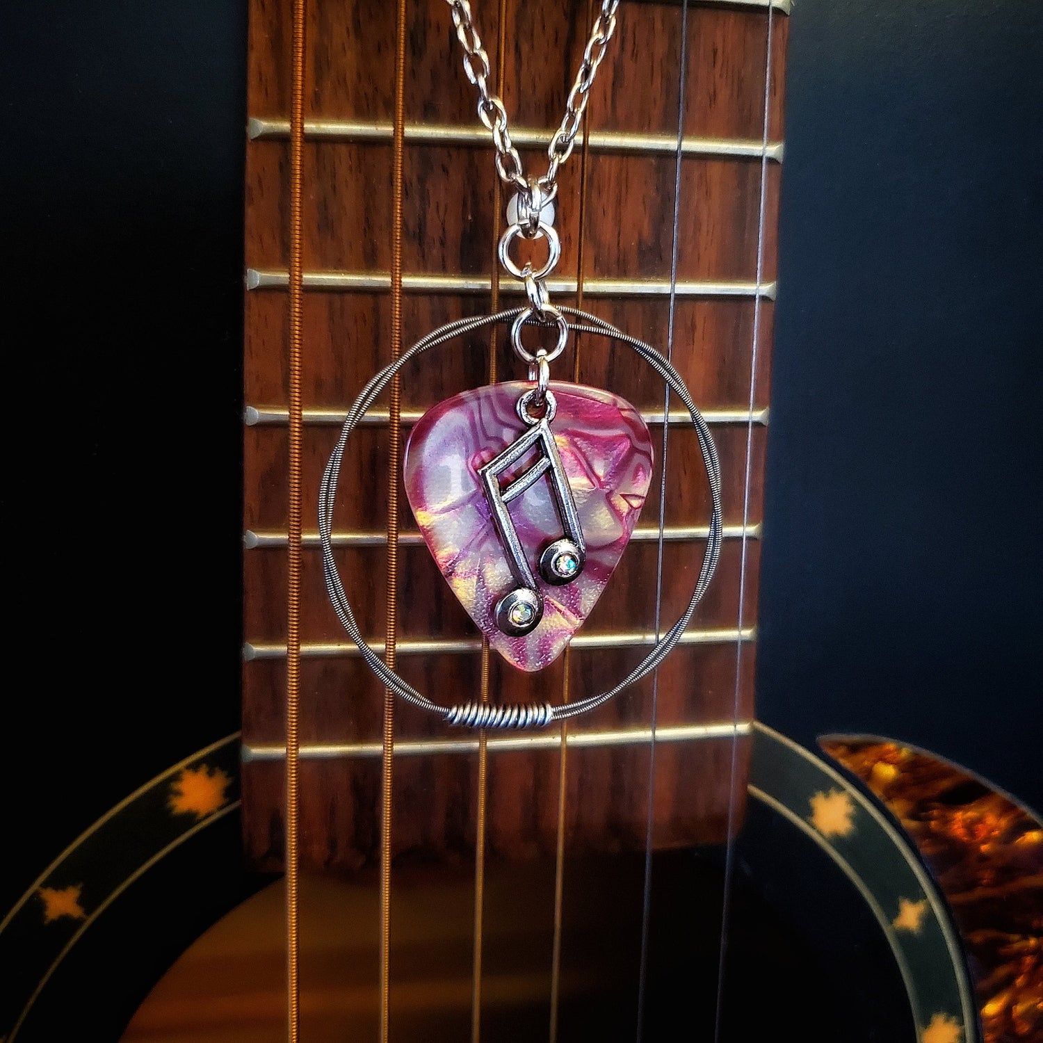 A necklace made from a red marbled guitar pick and upcycled guitar strings with a silver coloured chain hangs in front of a black guitar