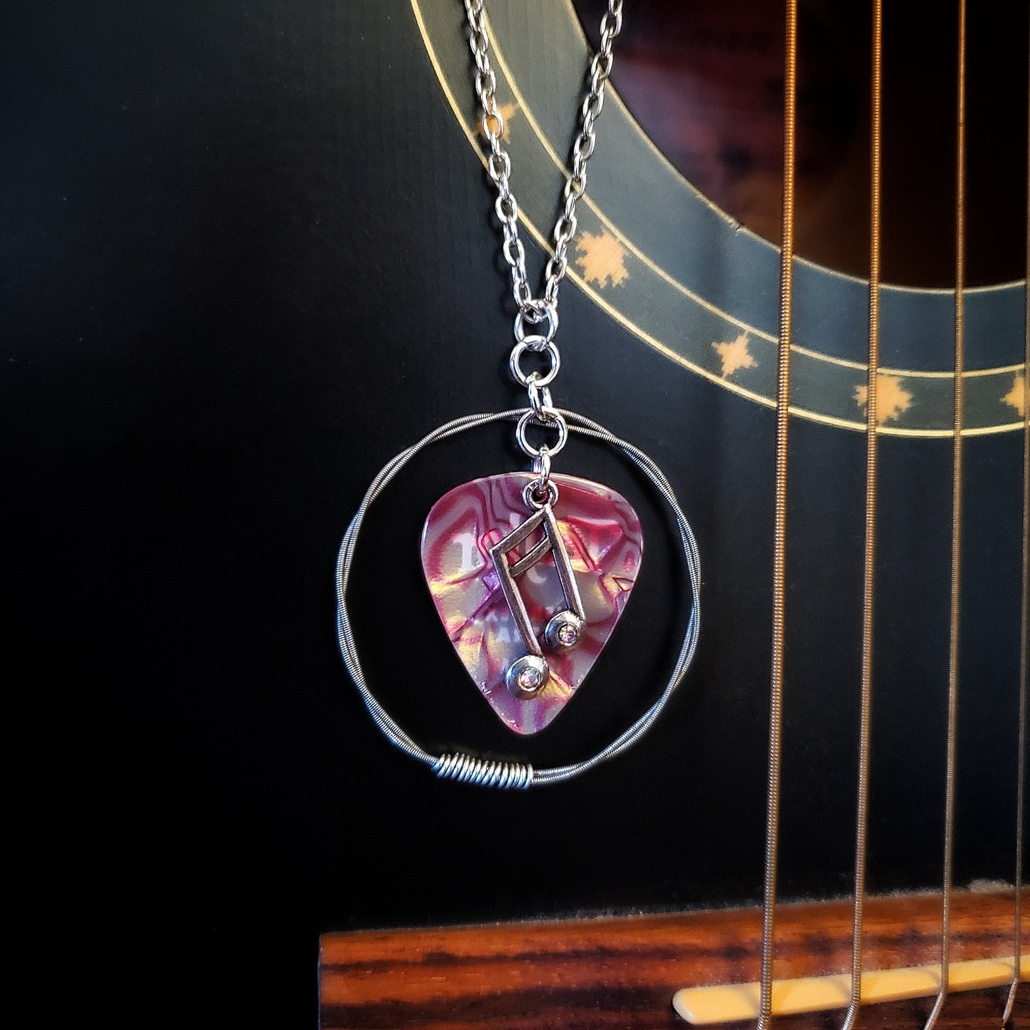 A necklace made from a red marbled guitar pick and upcycled guitar strings with a silver coloured chain hangs in front of a black guitar