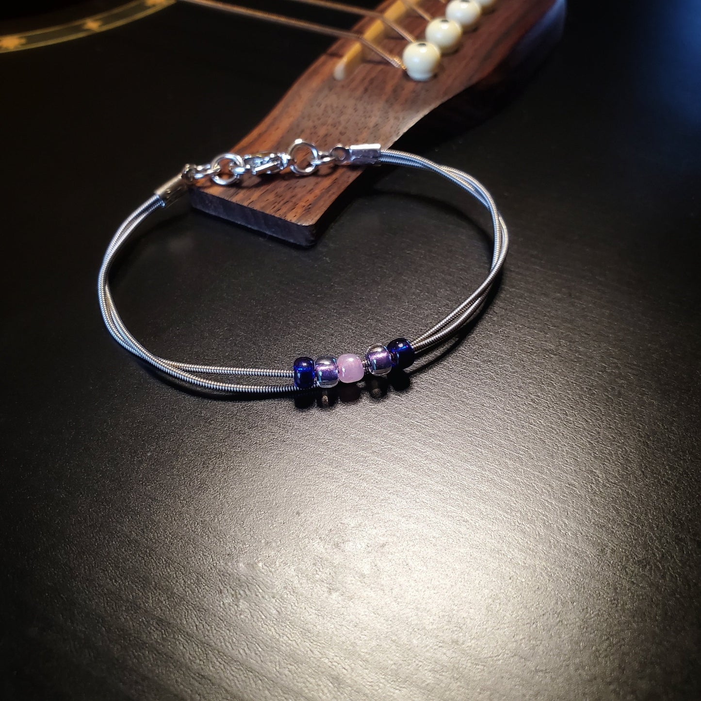 guitar string bracelet with blue, purple and pink bead representing the bisexual pride flag lying on a black guitar