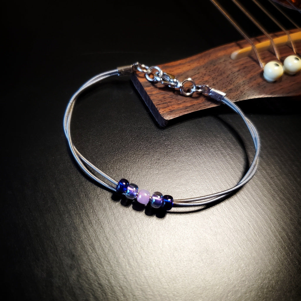 guitar string bracelet with blue, purple and pink bead representing the bisexual pride flag lying on a black guitar