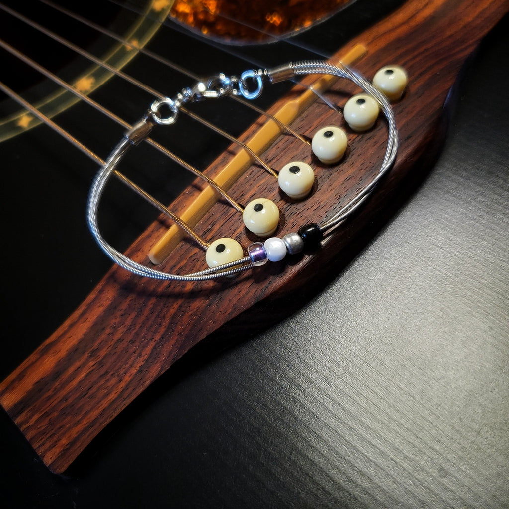 silver coloures clasp style bracelet with 4 glass beads representing the asexual pride flag (black, grey, white & purple) -bracelet is on a black guitar