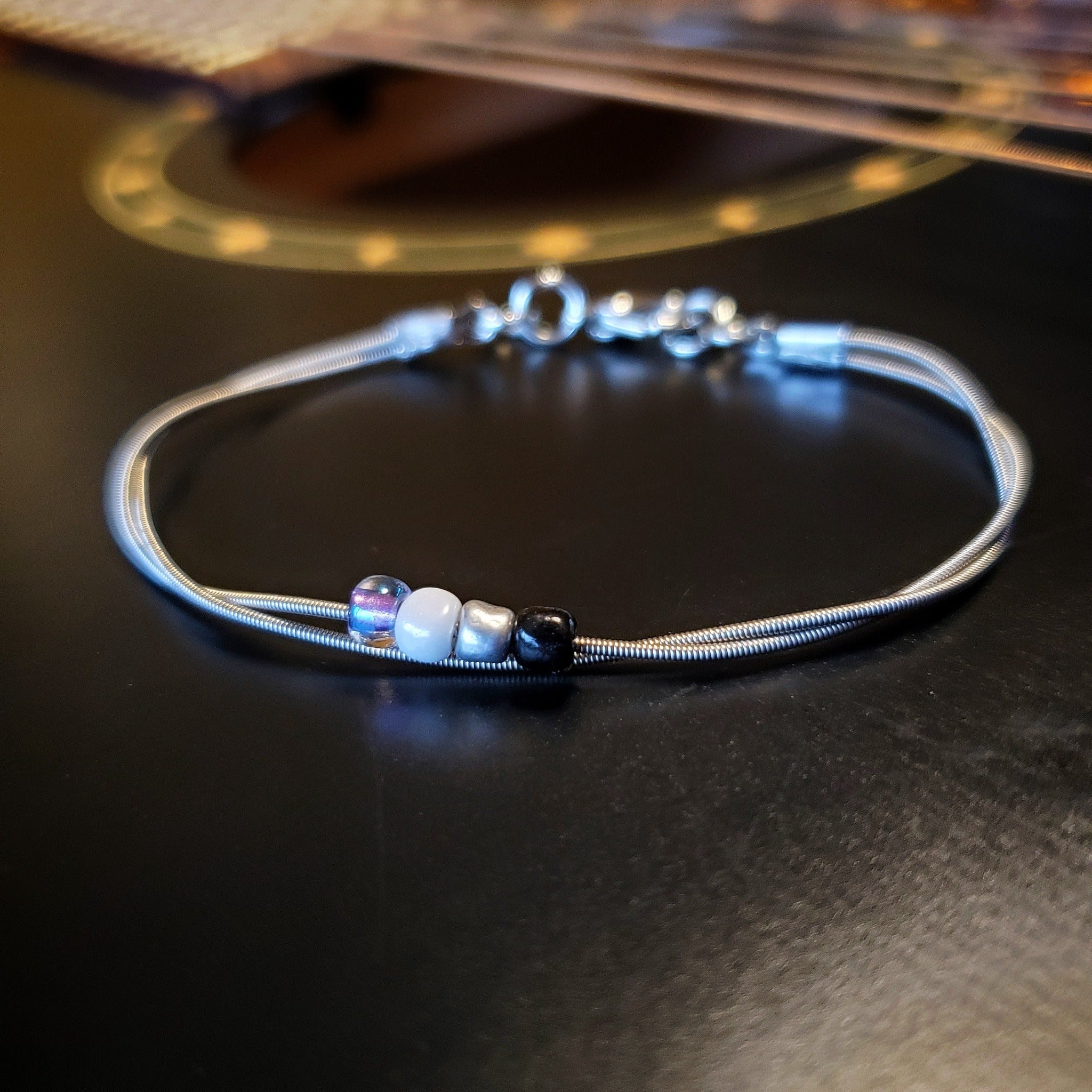 silver coloures clasp style bracelet with 4 glass beads representing the asexual pride flag (black, grey, white & purple)  -bracelet is on a black guitar