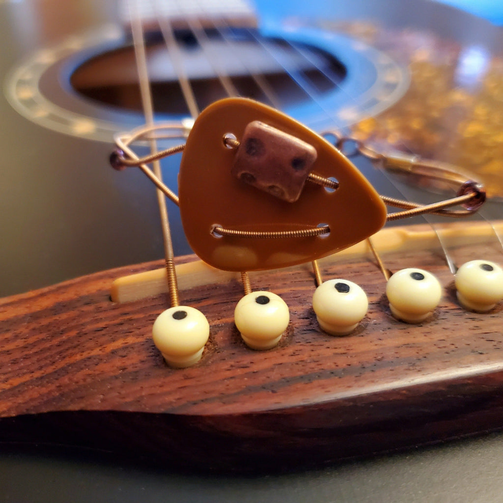 Bracelet made from a brown plectrum and upcycled copper coloured guitar strings -bracelets is sitting on the bridge of a black guitar 