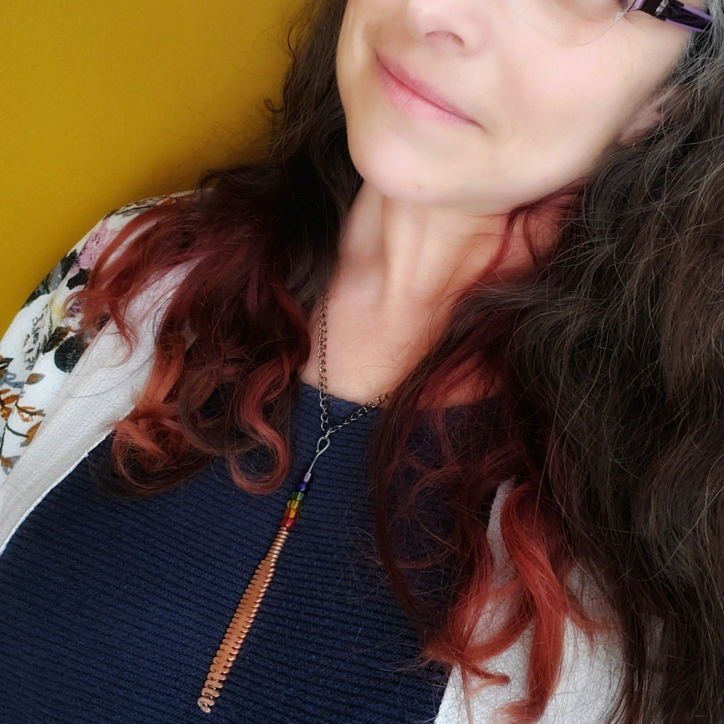 Woman with long dark hair and glasses wearing a hammered piano string pendant with 6 LGBTQ flag coloured beads with chain