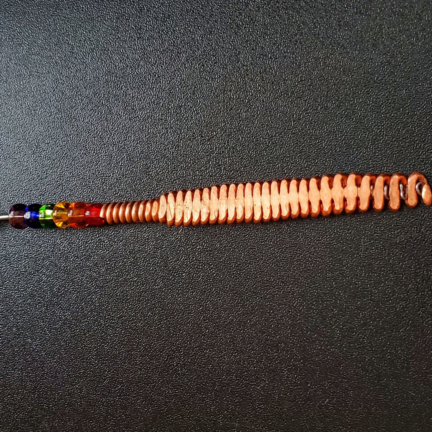 Hammered piano string pendant with 6 LGBTQ flag coloured beads with chain on a piano black background 
