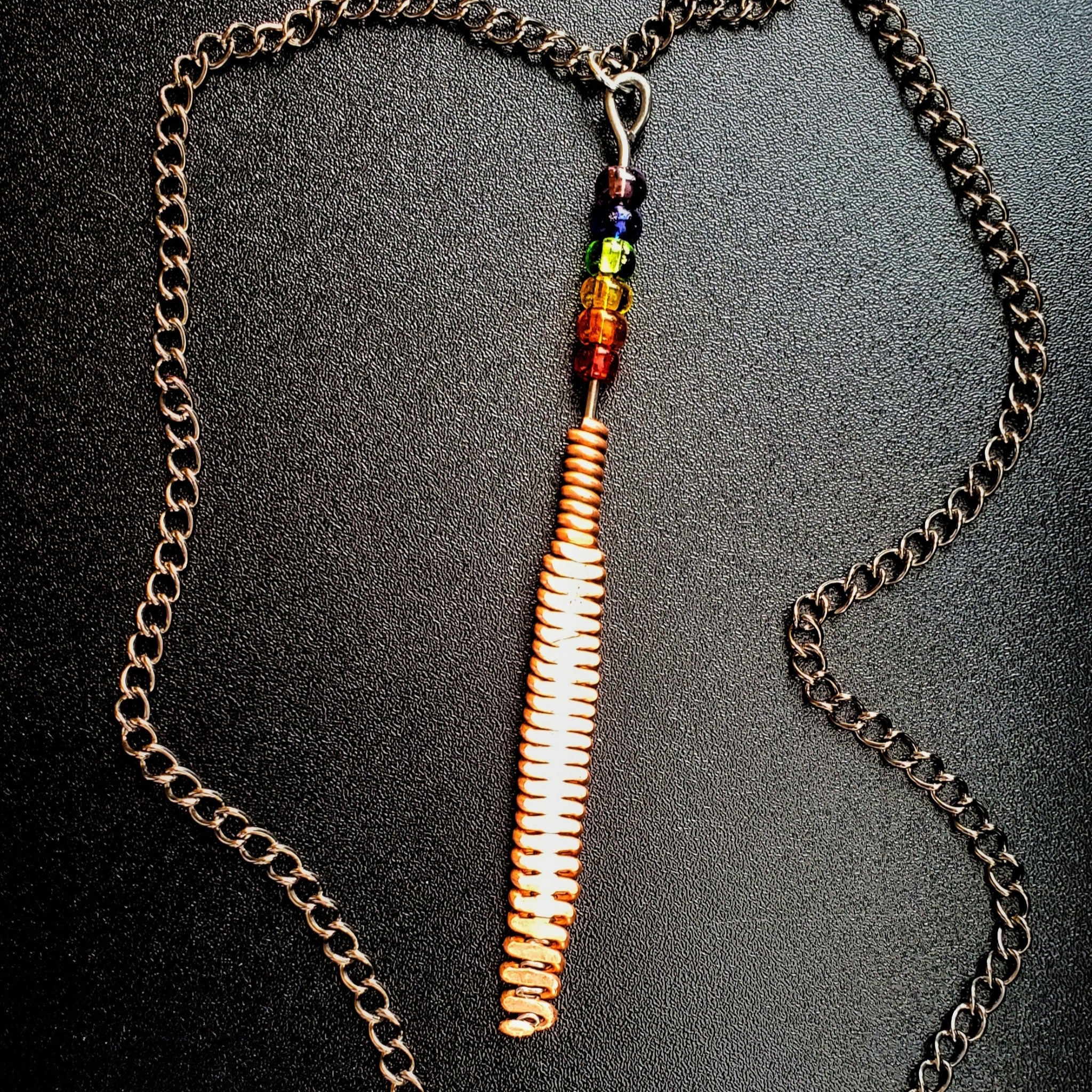 Hammered piano string pendant with 6 LGBTQ flag coloured beads with chain on a piano black background 