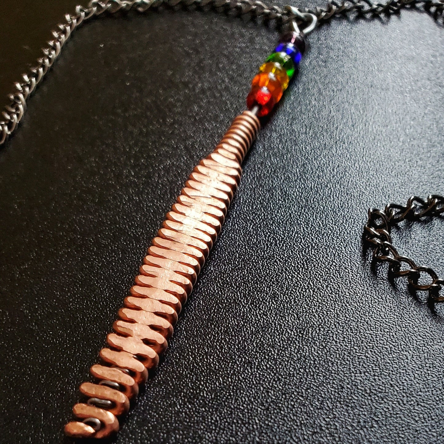 Hammered piano string pendant with 6 LGBTQ flag coloured beads with chain on a piano black background 