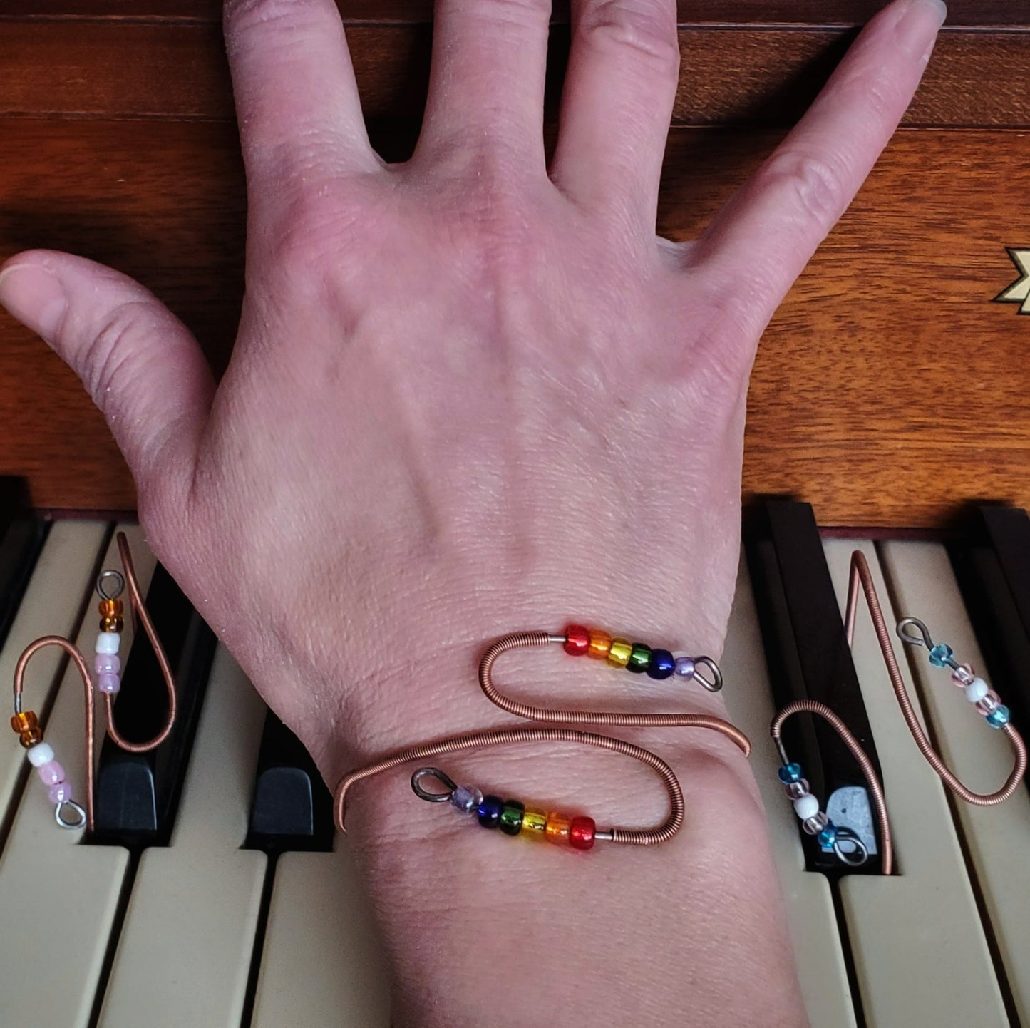 wrist with a cuff style bracelet made from upcycled piano string with beads representing the LGBTQ pride flag - wrist and 2 other bracelets sitting on piano keys