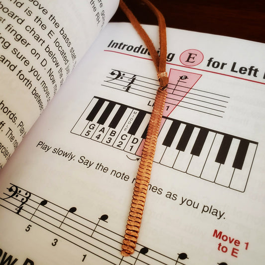 bookmark made from a hammered piano string and a piece of rust coloured suede sitting on the page of a book with music notes and words written in black on white background
