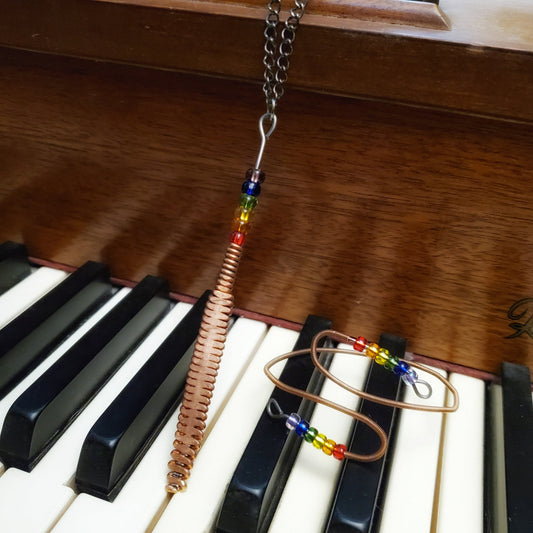 necklace and bracelet made from upcycled piano strings with beads representing the LGBTQ pride flag, sitting on piano keys
