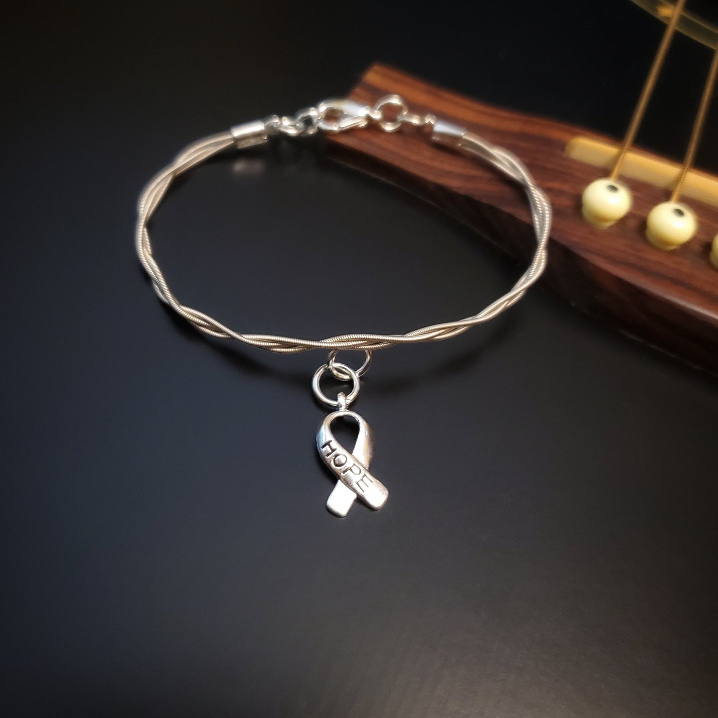 braided guitar string clasp style bracelet with ribbon style charm the word "HOPE" is engraved on the charm - the bracelet is lying on the bridge of a black guitar