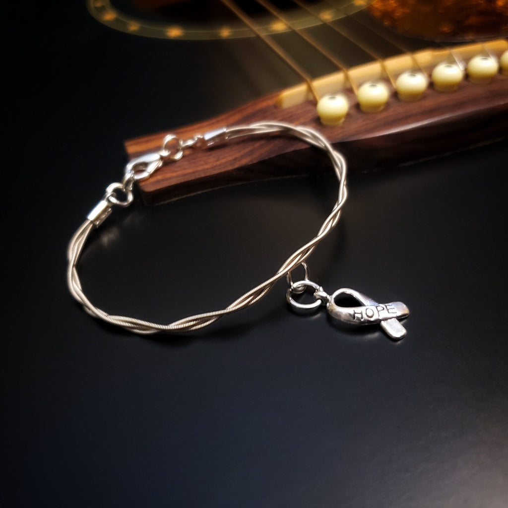 braided guitar string clasp style bracelet with ribbon style charm the word "HOPE" is engraved on the charm - the bracelet is lying on the bridge of a black guitar