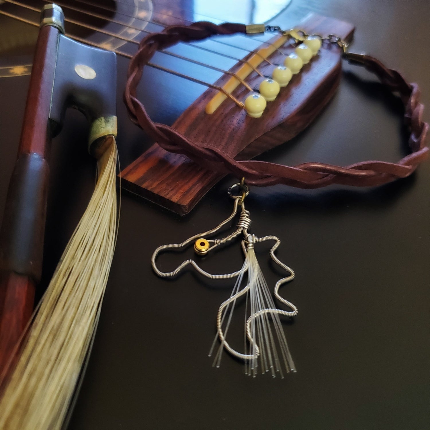 pendant made from an upcycled guitar string and genuine horse hair from a violin bow hangs from a braided suede necklace - the necklace is next to a violin bow and is lying on a black guitar
