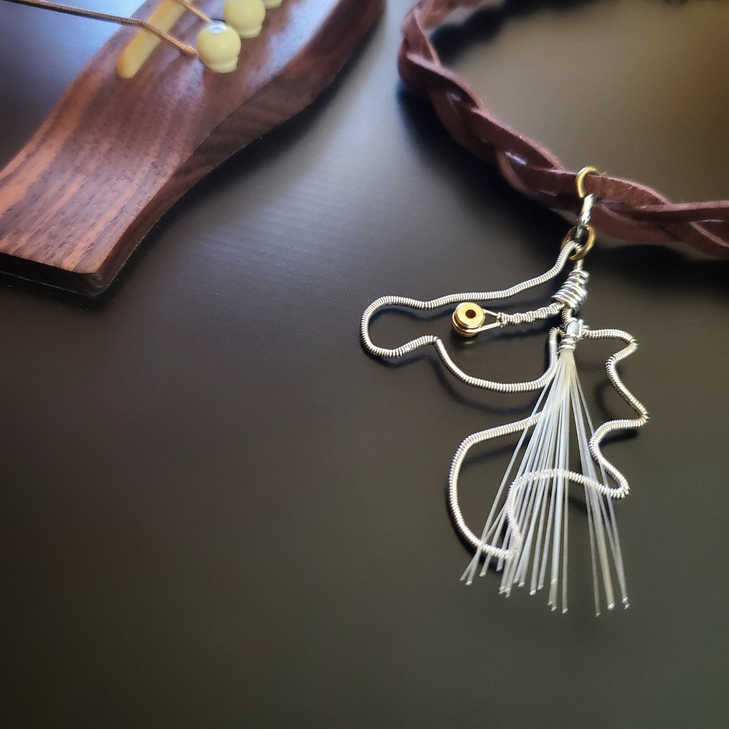 pendant made from an upcycled guitar string and genuine horse hair from a violin bow hangs from a braided suede necklace - the necklace is lying on a black guitar
