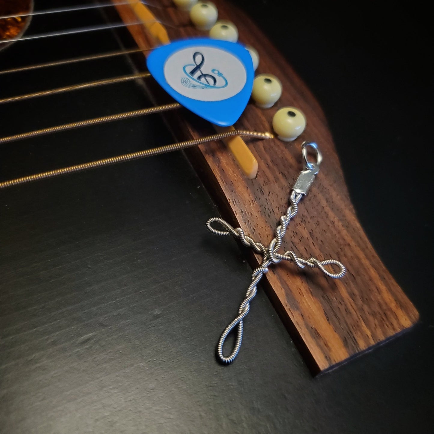 Upcycled guitar string cross style pendant lying on the bridge of a guitar next to a blue guitar pick