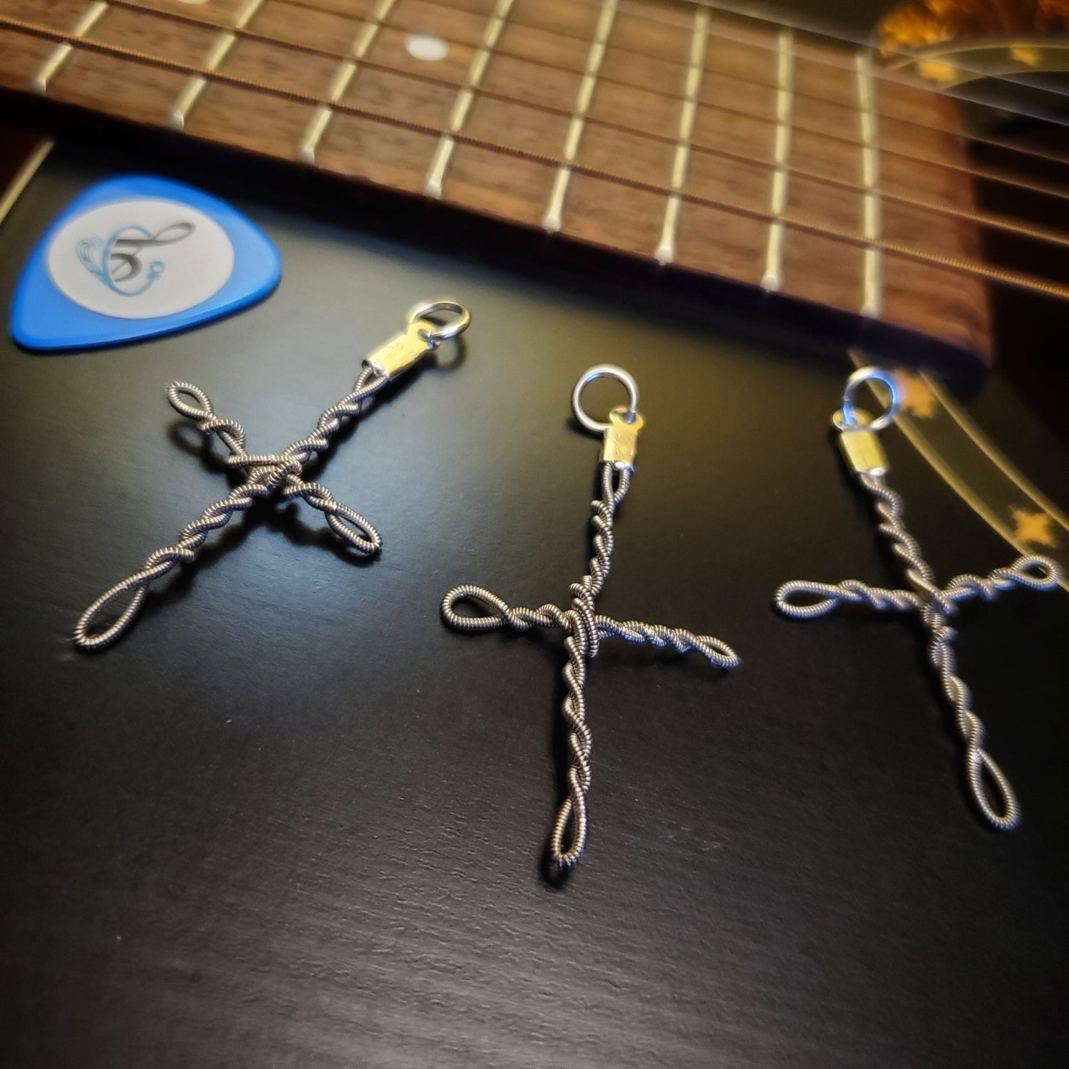 Three upcycled guitar string cross style pendants lying on the bridge of a guitar next to a blue guitar pick