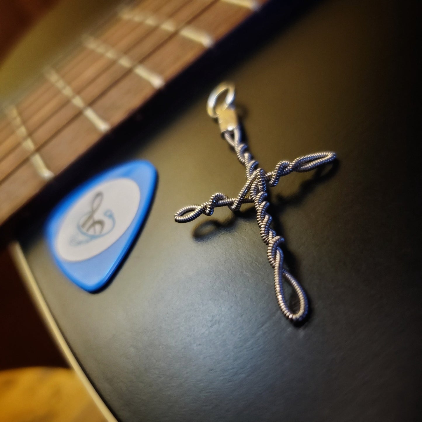 Upcycled guitar string cross style pendant lying next to the bridge of a guitar next to a blue guitar pick