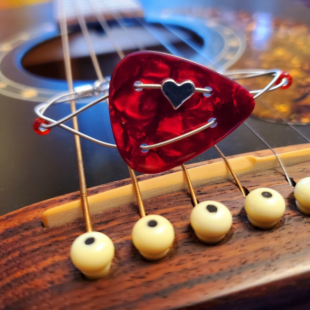 Bracelet made with a red guitar pick and upcycled guitar string with a small silver coloured heart sits on the bridge of a black guitar.