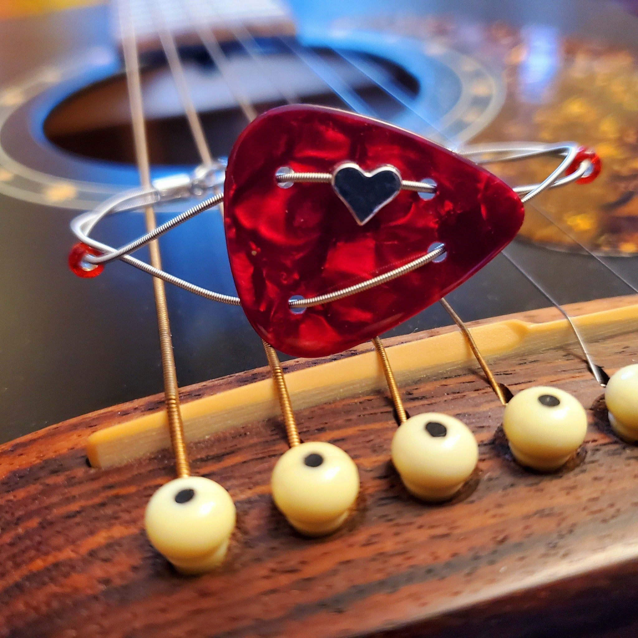 Bracelet made with a red guitar pick and upcycled guitar string with a small silver coloured heart sits on the bridge of a black guitar.