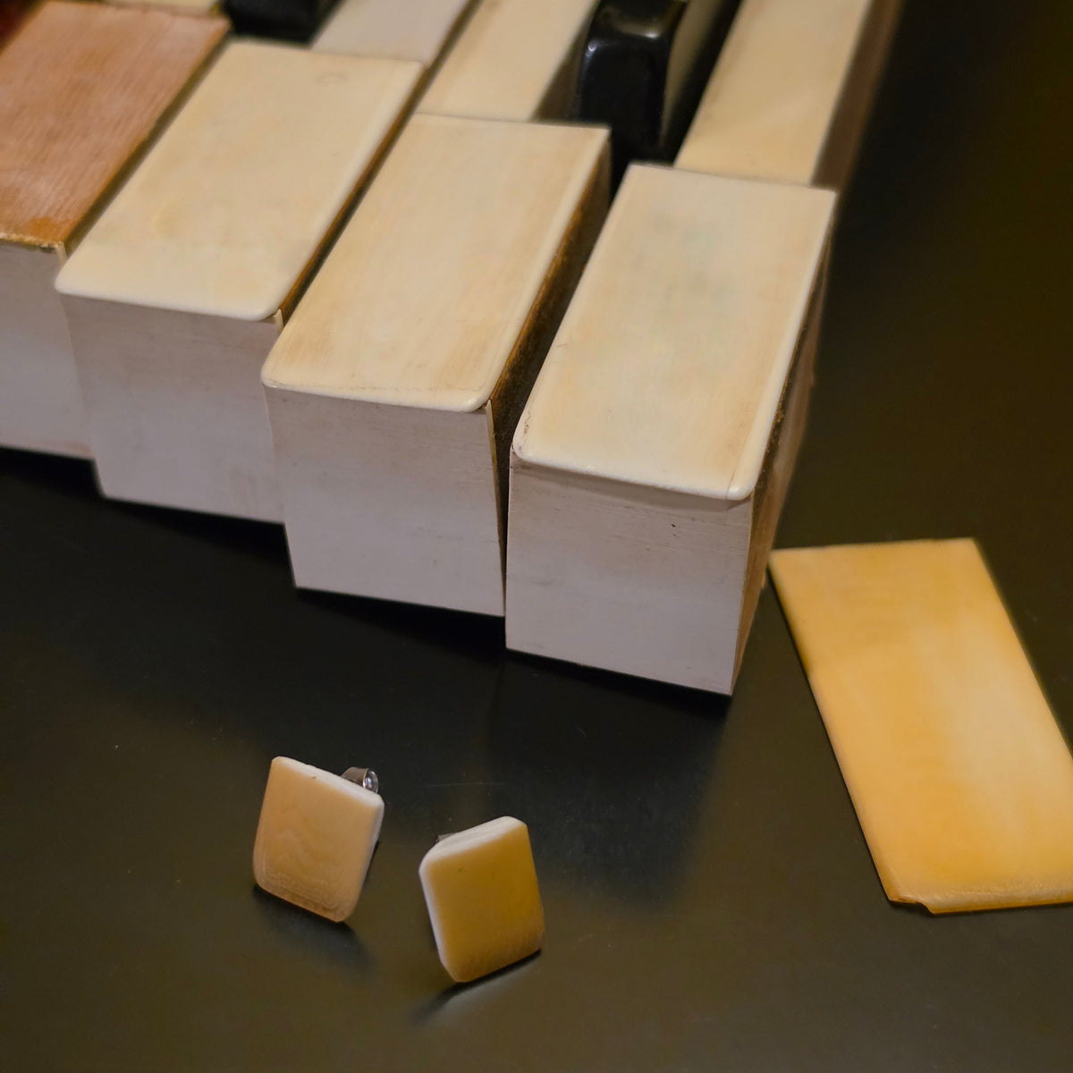 Close-Up of A pair of square piano ivory earrings next to piano keys and a piece of piano ivory