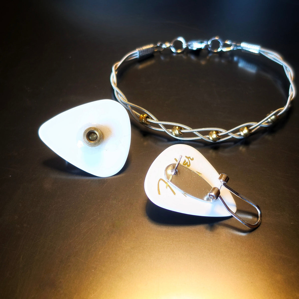 Closeup of Braided Guitar String and Gold Coloured Ballend Clasp Style Bracelet and Guitar Pick and Ballend Earrings Jewelry Set on Dark Background 