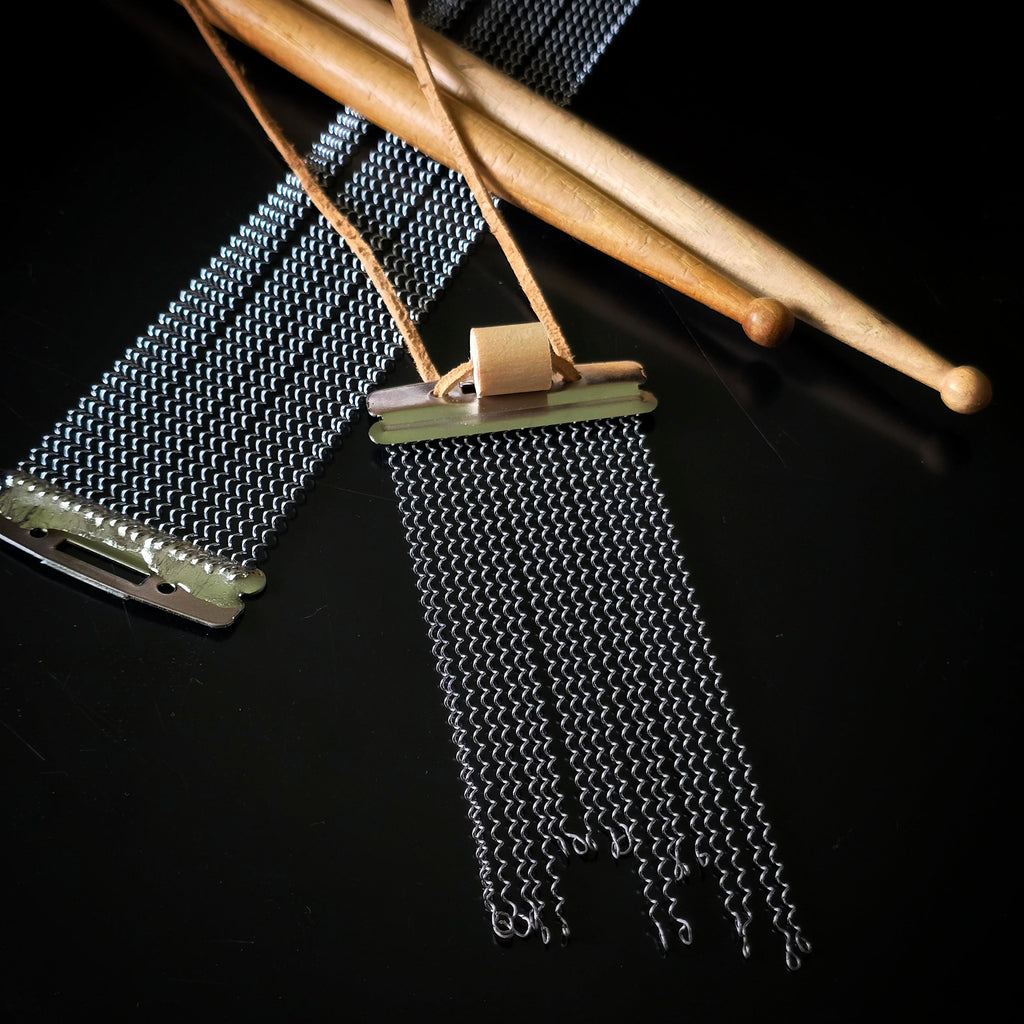 A closeup of a snare drum string fringe necklace with light brown suede straps (with a drumstick bead), displayed on a black surface with wooden drumsticks and snare drum strings the background.