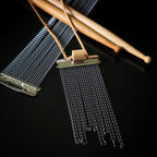 A closeup of a snare drum string fringe necklace with light brown suede straps (with a drumstick bead), displayed on a black surface with wooden drumsticks and snare drum strings the background.