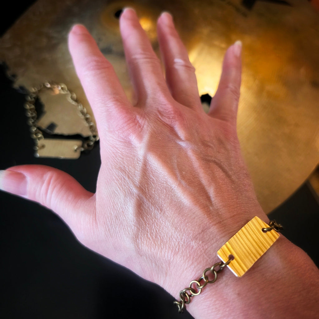 Closeup of a person's wrist wearing a gold-toned cymbal piece bracelet with chain links , against a dark background with a blurred brass cymbal.