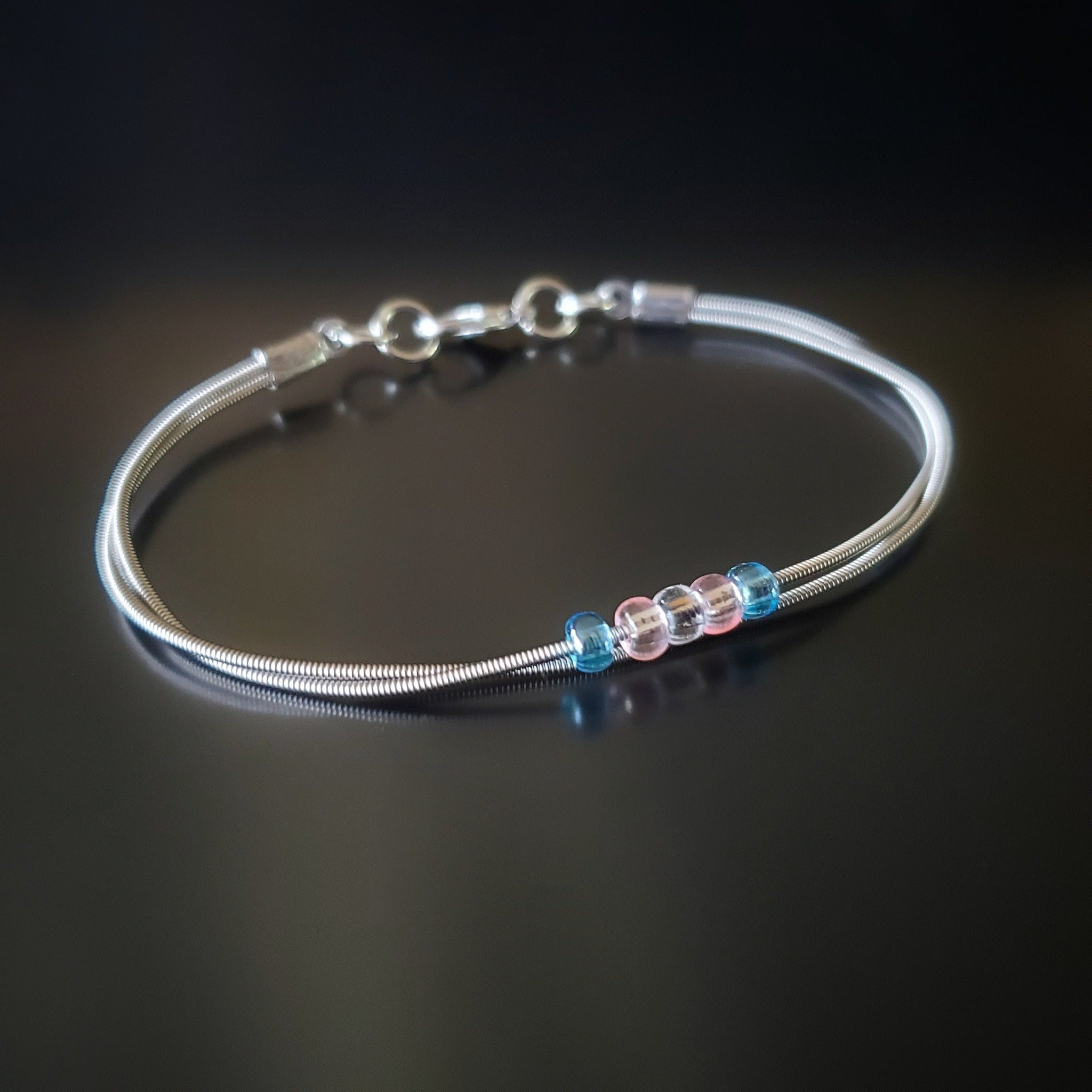 silver coloured clasp style bracelet made from upcycled guitar strings - there are 5 beads representing the colours of the transgender pride flag - 2 blue, 2 pink and 1 white